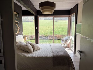 Une chambre avec un grand lit, une chaise et une vue sur un champ verdoyant, visible à travers une porte coulissante.
