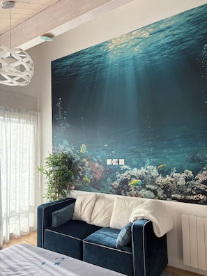 A room with a large wall mural of an underwater scene, a blue sofa with cushions, a potted plant, and a white radiator. - Architect-designed home with a heated pool, just a stone's throw from Geneva and the ski slopes (Présilly)