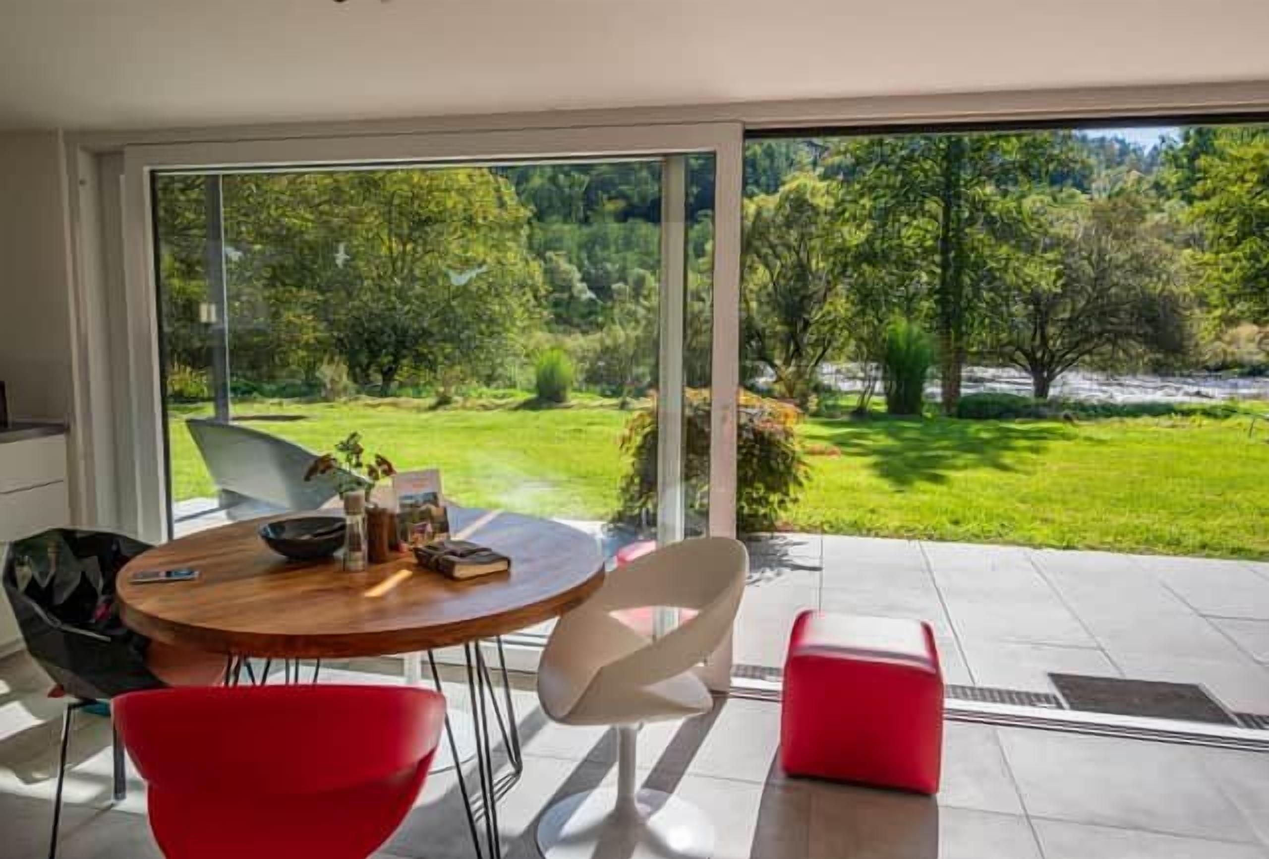 A modern dining area with a table and chairs, a red bench, and a view of a lush garden through large glass doors.