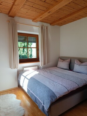 Ein Schlafzimmer mit Holzboden, einem großen Fenster mit Vorhängen, einem Bett mit gemusterten Betttextilien und einem weißen, flauschigen Teppich.