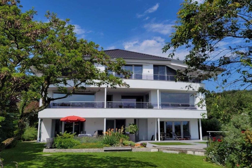 A two-story modern house with a balcony, a red umbrella, and a well-maintained garden.
