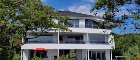 A two-story modern house with a balcony, a red umbrella, and a well-maintained garden.