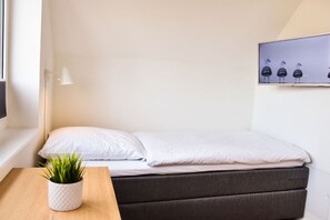A single bed with a grey mattress and white bedding, a bedside table with a plant, a wall-mounted TV, and a white lamp. - Holiday apartment in Büsum with swimming pool (Büsum)