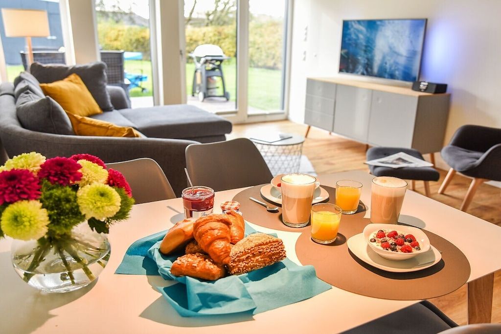 Une table est dressée dans un salon moderne avec un canapé et un téléviseur, et elle est garnie de viennoiseries, d’un saladier de fruits et de diverses boissons pour le petit-déjeuner.