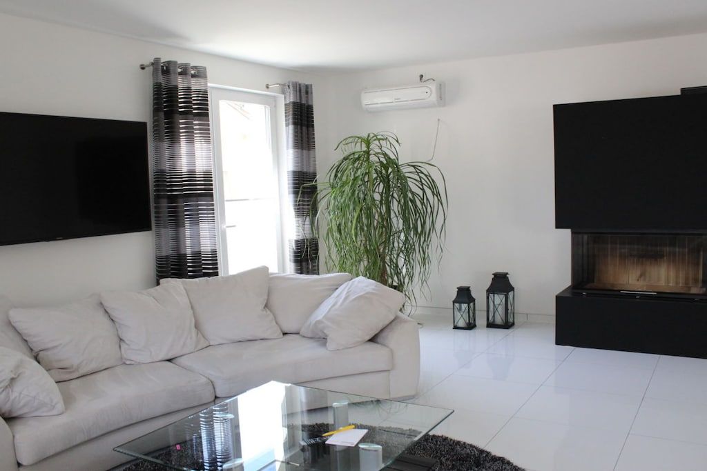 Een moderne woonkamer met een witte bank, een glazen salontafel, een grote plant, een open haard en een tv aan de muur.