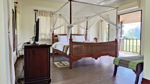 Een hemelbed met baldakijn, een televisie op een houten standaard, en een balkon met uitzicht op groen.