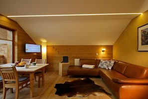 Ein gemütliches Zimmer mit Holzbalkendecke, einem braunen Ledersofa, einem Esstisch mit Stühlen und einem angebrachten Flachbildfernseher.