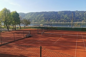 A tennis court with a net, surrounded by trees and a view of a lake and hills. - Seehäuser Ramsbacher Haus 2 (Sattendorf)