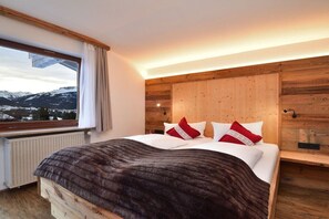 A bedroom with a wooden headboard, a large window with a view of snow-capped mountains, and a bed with white bedding and red pillows. - Apartment Widderstein (Riezlern)