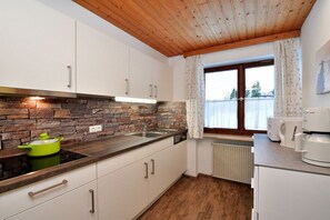 A kitchen with white cabinets, a stone backsplash, a wooden ceiling, a window with curtains, and a green pot on the stove. - Apartment Widderstein (Riezlern)