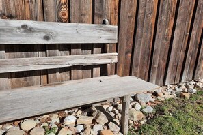 Eine Holzbank mit querverlaufender Lehne und flacher Sitzfläche, vor einem Holzzaun platziert und von einem Kiesweg umgeben.