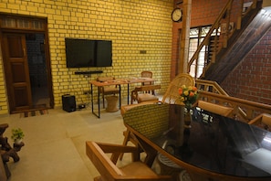A room with a brick wall, a flat-screen TV mounted above a wooden table, a staircase with wooden railings, and a wicker chair with a vase of flowers. - Relaxing Holiday Villa in Santhapuram, Kanyakumari (Nagercoil)