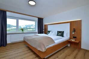 A modern bedroom with a wooden bed, a nightstand, a bedside lamp, and a window with a view of the outdoors. - Holiday apartment with 3 bedrooms (Riezlern)