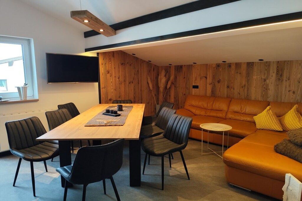 A modern living room with a wooden wall, a sofa, a dining table with chairs, and a flat-screen TV mounted on the wall.