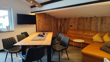 A modern living room with a wooden wall, a sofa, a dining table with chairs, and a flat-screen TV mounted on the wall.