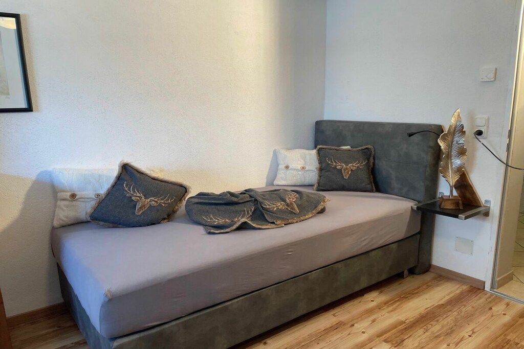 A modern bedroom with a grey sofa bed, decorative pillows, a wooden floor, and a wall-mounted shelf.