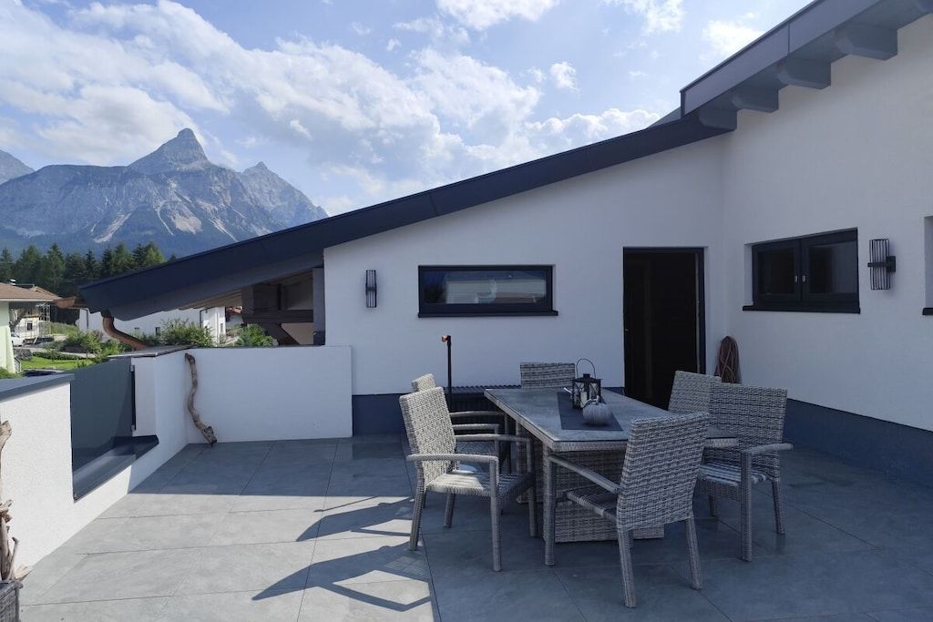 An outdoor patio with a dining table and chairs, a mountain view, and a modern building with large windows.