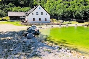 Ein weißes Gebäude mit geneigtem Dach, einer kleinen Veranda und einem Teich mit grünem Wasser.
