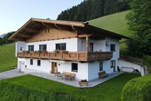 Un chalet de dos pisos con balcón, vigas de madera y techo inclinado, rodeado de exuberante vegetación.
