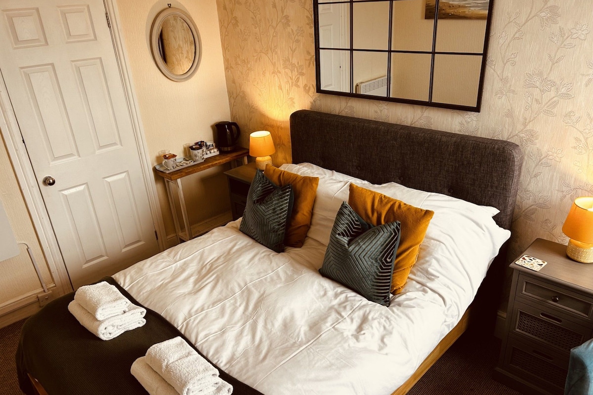 A neatly made bed with pillows and folded towels, a bedside table with a lamp, and a wall-mounted mirror.