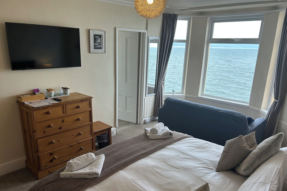 A hotel room with a bed, a dresser, a flat-screen TV, and a view of the sea through large windows.