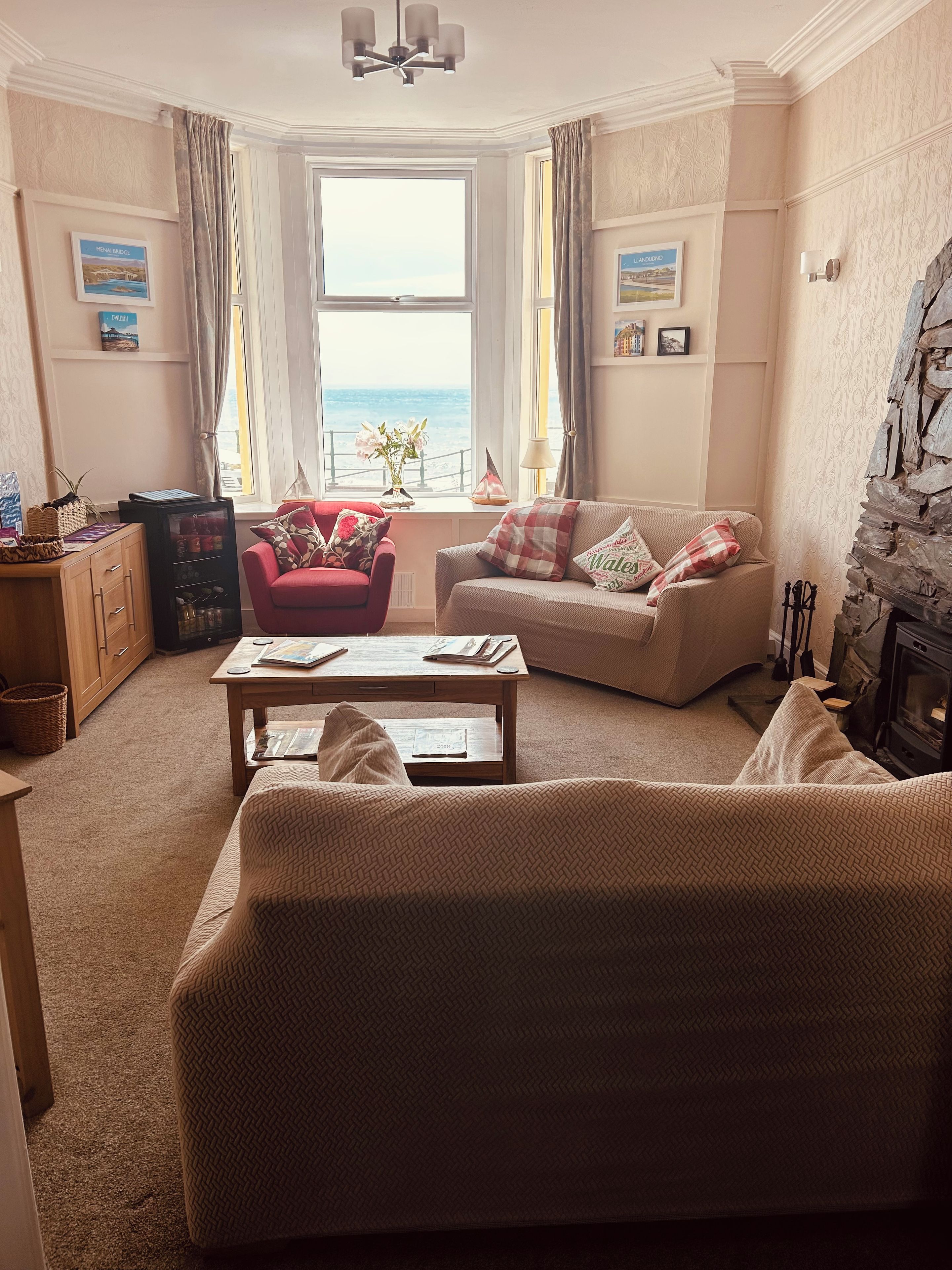 A cozy living room with a sofa, armchair, and a coffee table. There is a fireplace and a view of the sea through the window.