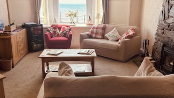 Una sala de estar acogedora con un sofá, un sillón y una mesa de centro. Hay una chimenea y vistas al mar a través de la ventana.