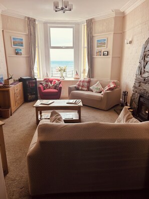 A cozy living room with a sofa, armchair, and a coffee table. There is a fireplace and a view of the sea through the window. - Awel Mor (Criccieth)