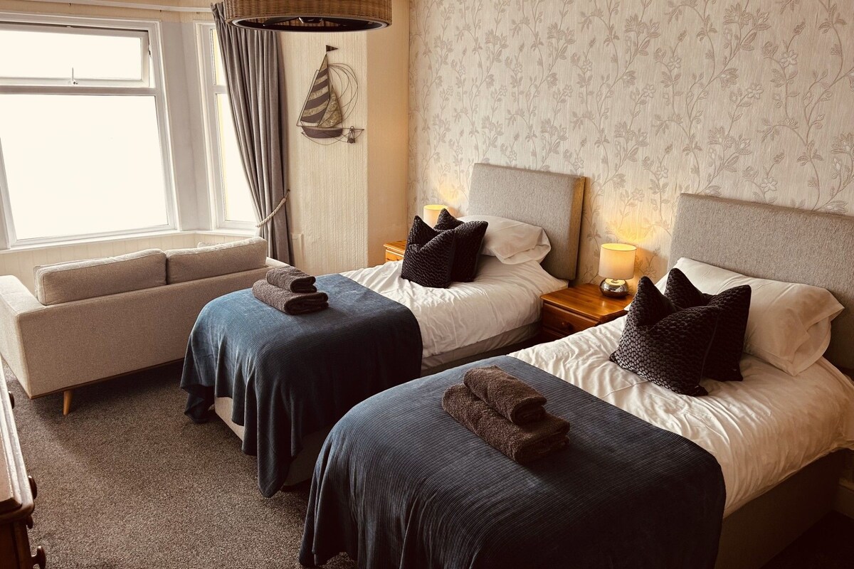 A hotel room with two beds, a sofa, a window with curtains, and a wall with floral wallpaper.