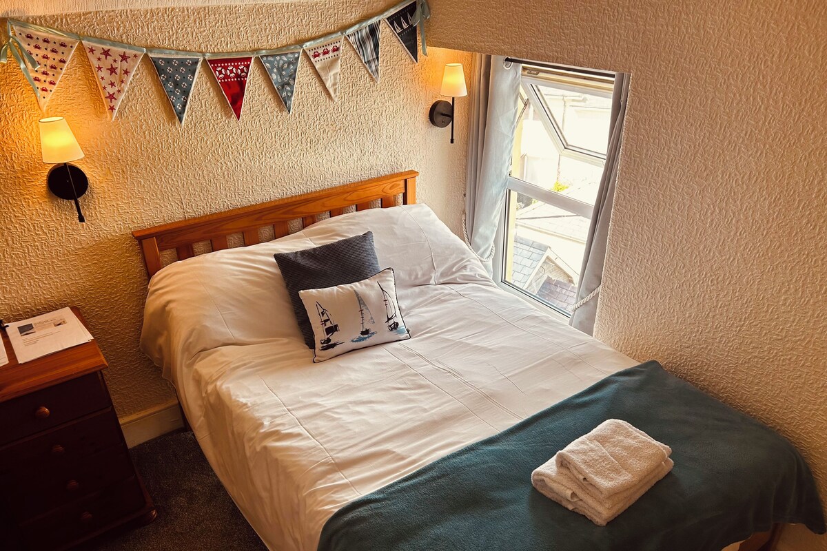 Una cama bien hecha con una almohada, una lámpara de noche y una ventana con cortinas.