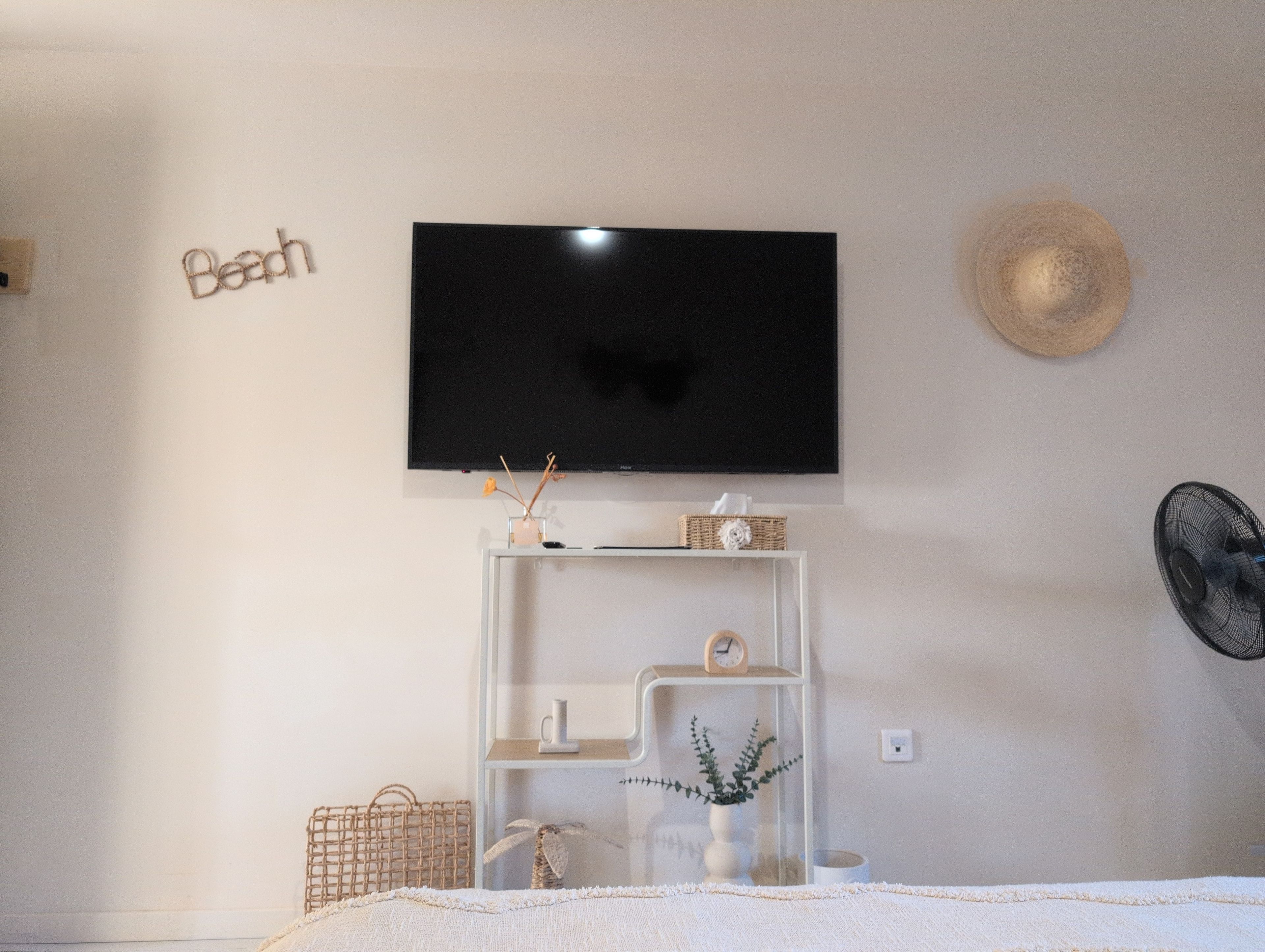 Un dormitorio con una televisión montada en la pared, una mesita de noche con un reloj, una cesta y una planta.