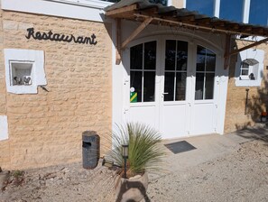A restaurant with a stone facade, a wooden pergola with string lights, and a small window with a rooster decoration.