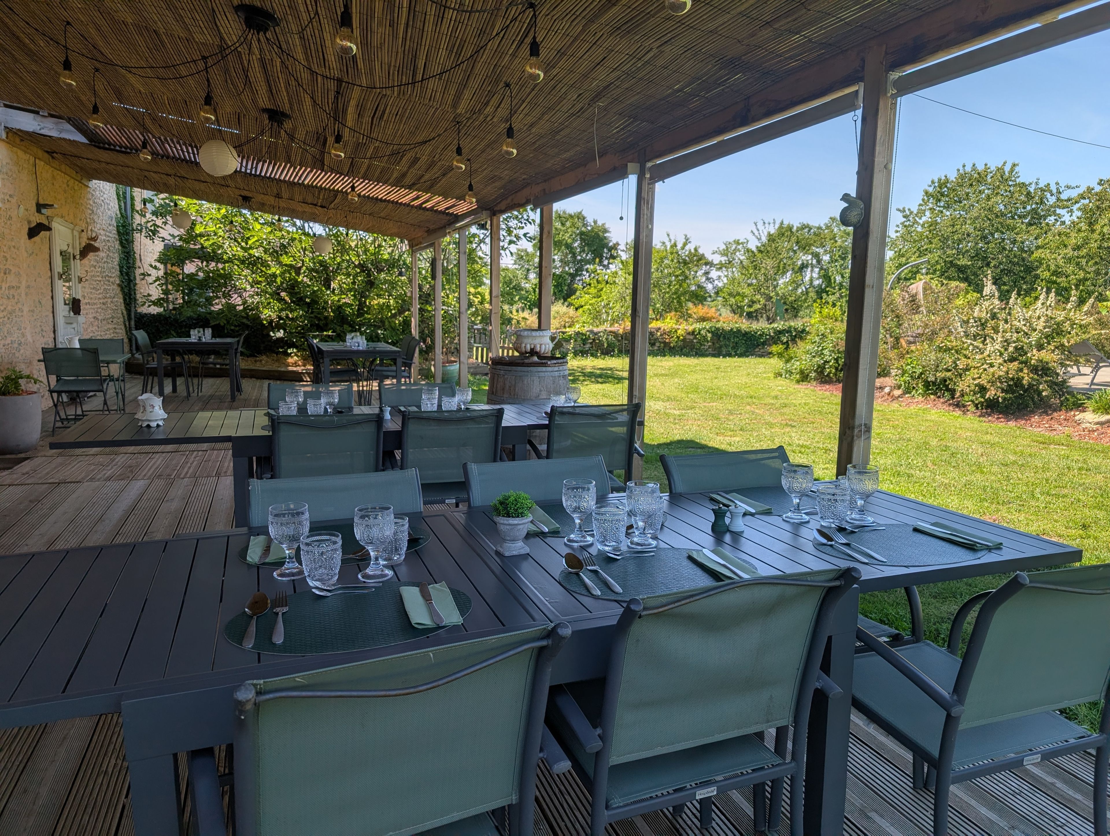 An outdoor dining area with tables set for four, surrounded by greenery and a clear sky.