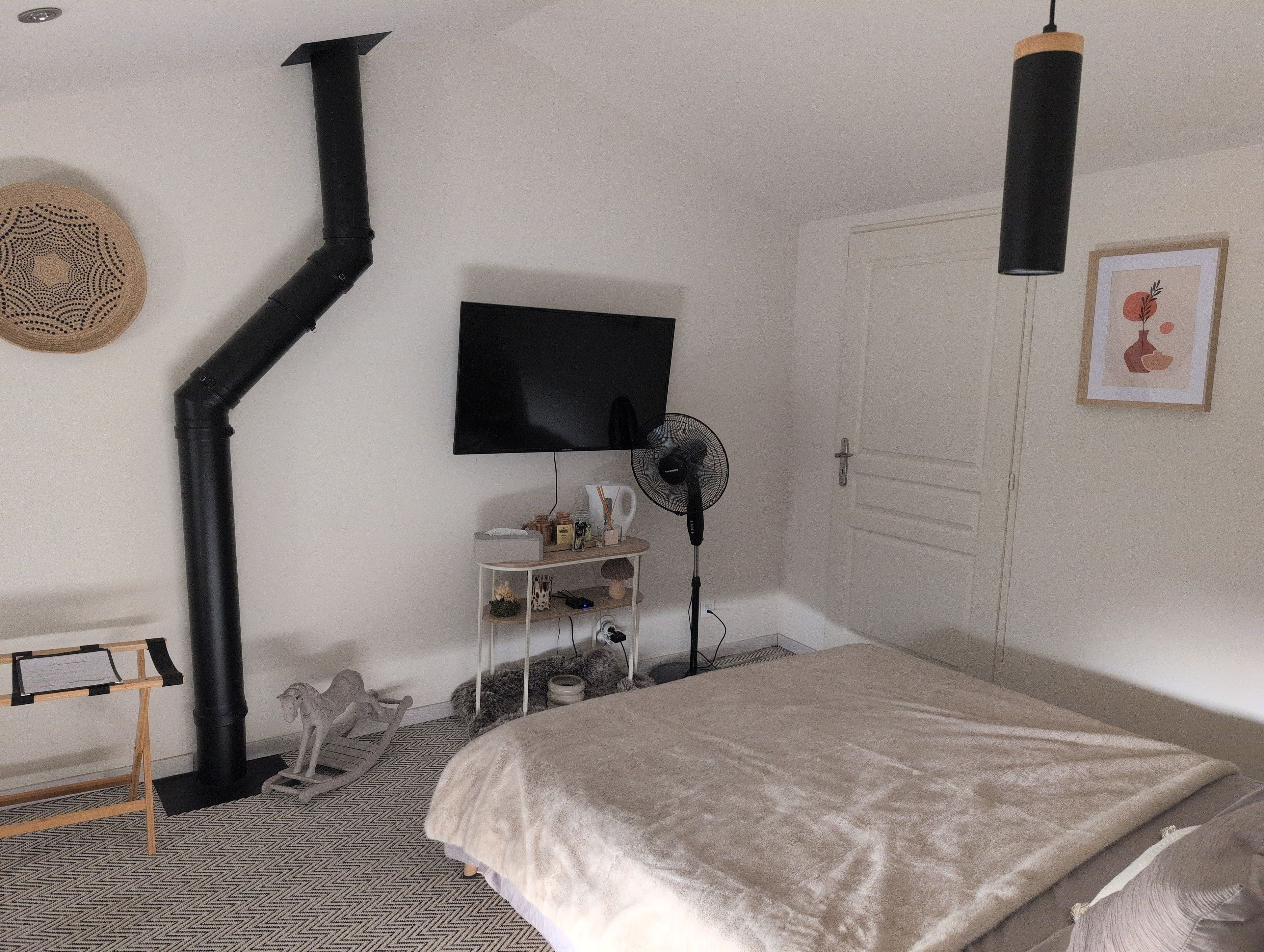 Een slaapkamer met een bed, een zwarte vloerventilator, een bevestigde televisie, een witte deur en een wandlamp.