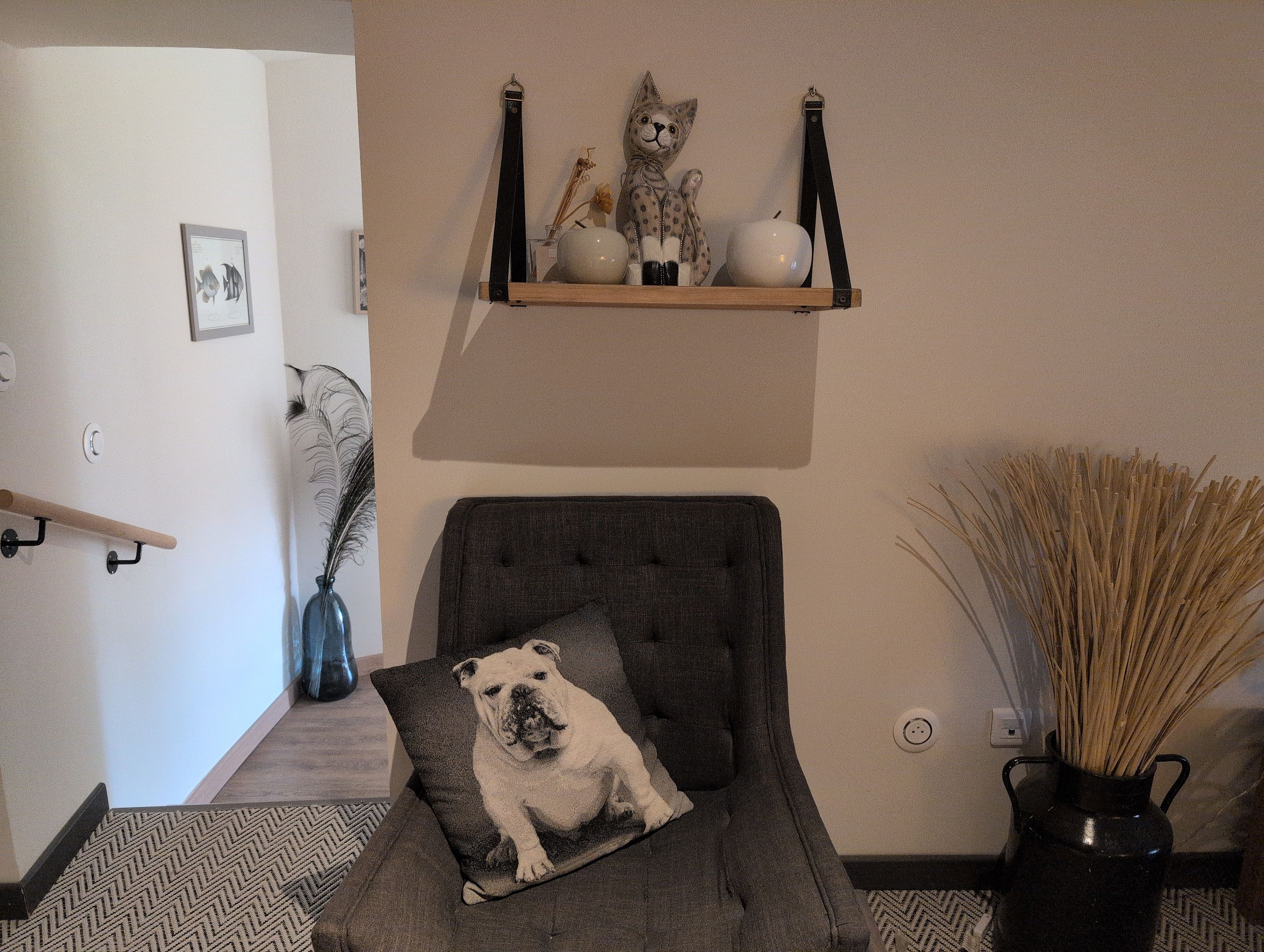Een woonkamer met een grijze fauteuil, een kussen met een bulldogafdruk, een houten plank met decoratieve objecten, een vaas met gedroogde planten, en een aan de muur hangend schilderij.