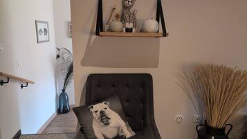 Een woonkamer met een grijze fauteuil, een kussen met een bulldogafdruk, een houten plank met decoratieve objecten, een vaas met gedroogde planten, en een aan de muur hangend schilderij.