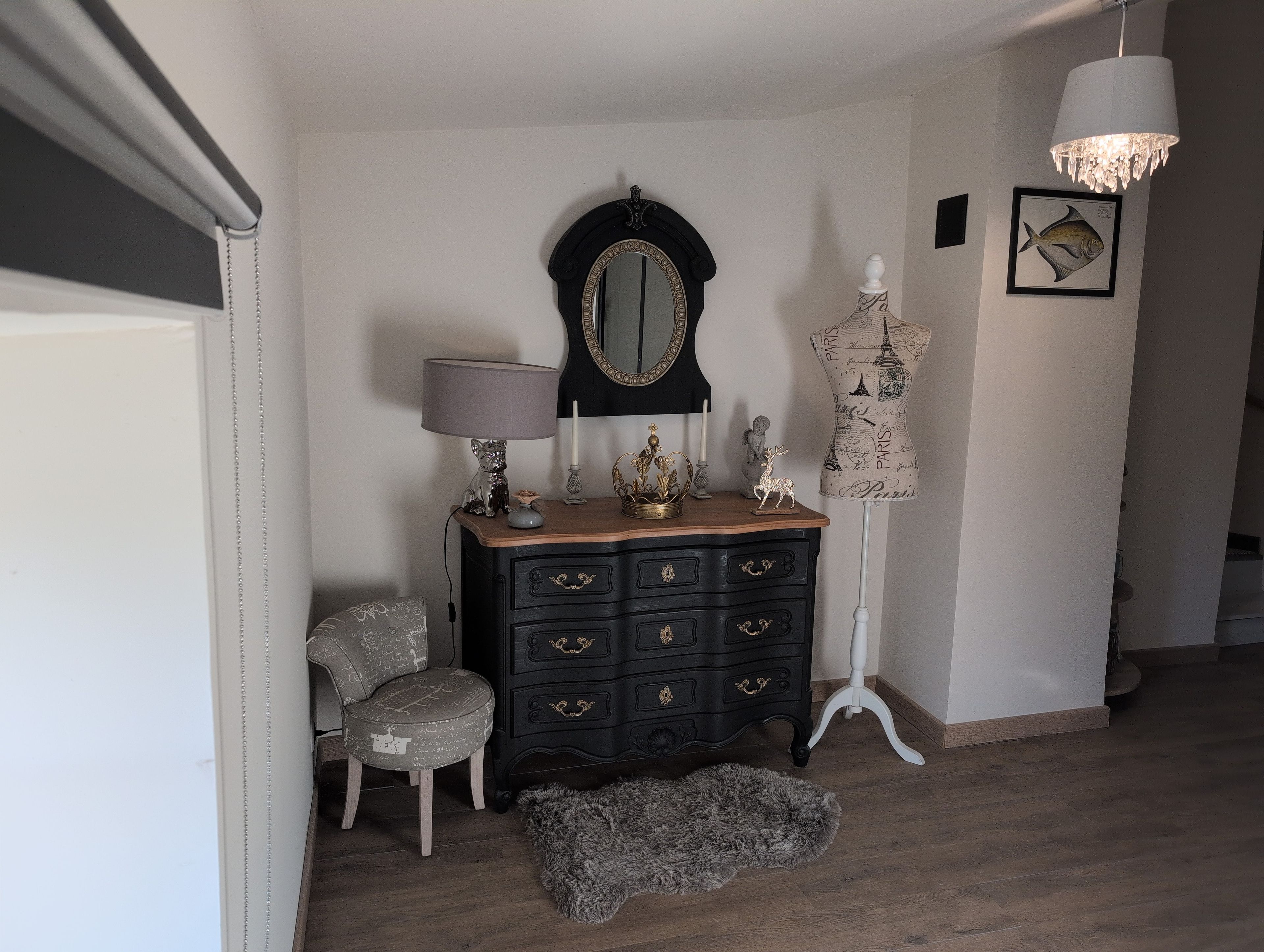 A room with a black dresser, a chair, a mirror, a mannequin, and a chandelier.