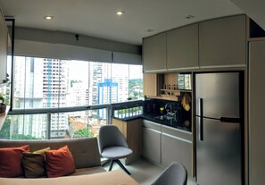 A modern kitchen with a stainless steel refrigerator, a dining area with a grey chair, and a large window offering a city view. - Studio - Berrini/ Faria Lima/ CGH Airport (São Paulo)