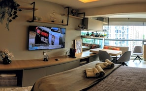 A hotel room with a bed, a TV displaying a news program, a desk with a computer, and a balcony with a view of buildings. - Studio - Berrini/ Faria Lima/ CGH Airport (São Paulo)