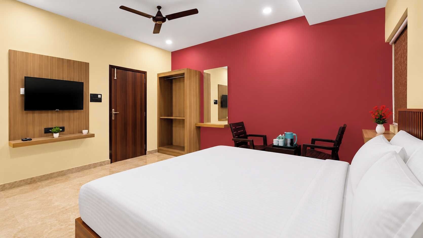 Habitación de hotel con cama, televisor montado en la pared, ventilador de techo, armario de madera, pared de acento roja y una mesita con un florero.