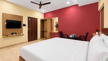 Habitación de hotel con cama, televisor montado en la pared, ventilador de techo, armario de madera, pared de acento roja y una mesita con un florero.