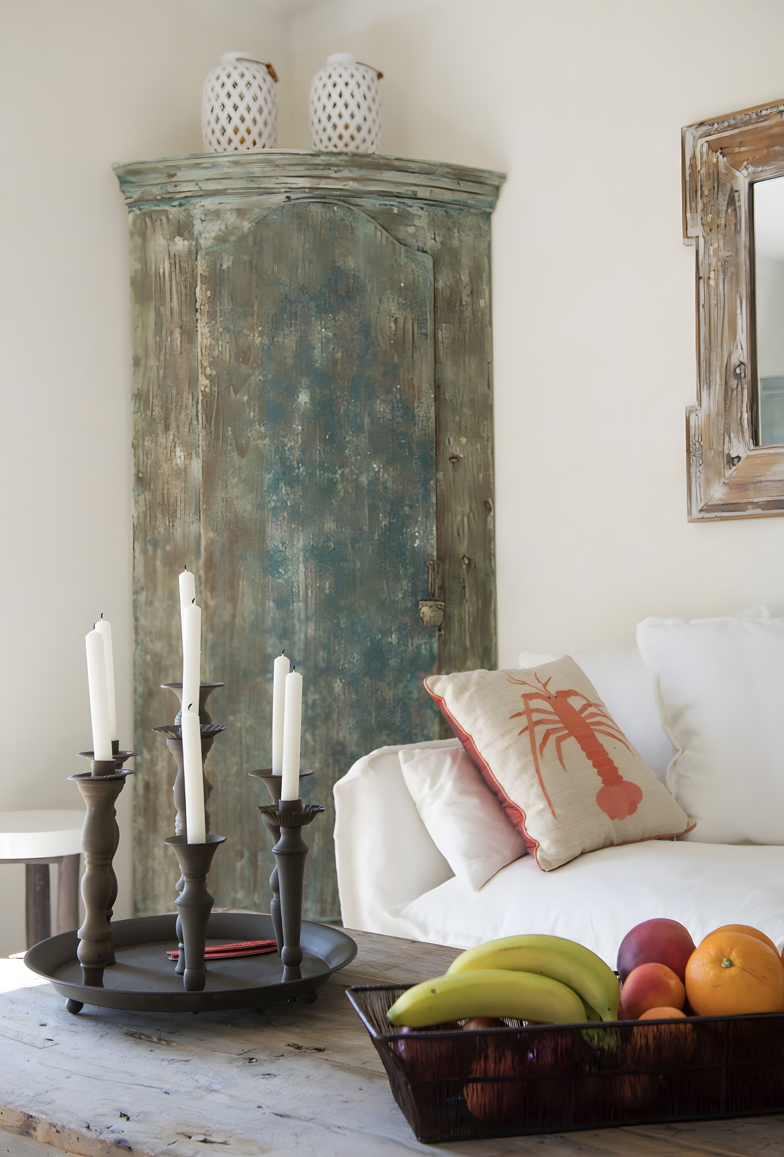 Un meuble en bois rustique, sur lequel sont posés deux bougies blanches sur un plateau, un panier de fruits contenant des bananes, des pommes et des oranges, et un canapé blanc agrémenté d’un coussin rouge et beige.