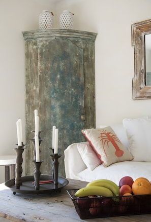 A rustic wooden cabinet with two white candles on a tray, a fruit bowl with bananas, apples, and oranges, and a white sofa with a red and beige pillow. - Villa 'La Bergerie' with Sea View, Wi-Fi and Air Conditioning (Coti-Chiavari)
