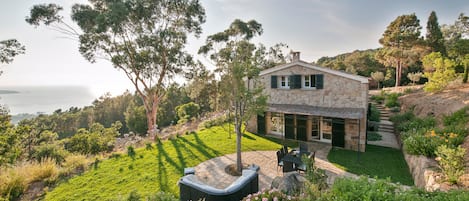 A stone house with a patio, surrounded by lush greenery and a view of the sea.