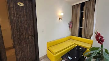 Una habitación con un sofá amarillo, una mesa negra, una puerta con un número e una planta.