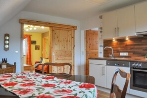 Eine kleine Küche mit Holzschränken, einem Esstisch mit blumigem Tischläufer und einer sichtbaren Uhr an der Wand.