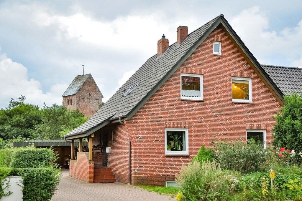 A red brick house with a gabled roof, a small attached structure, and a garden with various plants.