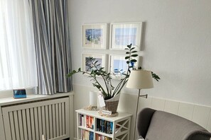 A room with a radiator, a bookshelf with books, a lamp, and a window with curtains. - Flurstraße - OG links (Wyk auf Föhr)