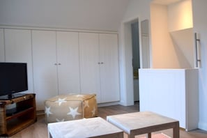 A room with a television on a wooden stand, a white wardrobe, a star-patterned ottoman, and a small wooden table. - Holiday home with 1 bedroom (Wyk auf Föhr)