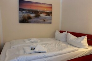 Une chambre d’hôtel avec un lit, du linge de lit blanc et un tableau représentant un coucher de soleil sur une plage.